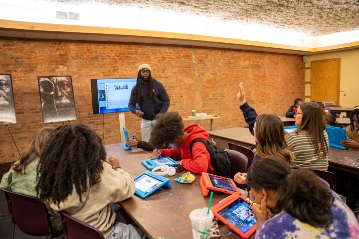 Students sit tables, looking down at iPads. A student raises her hand as the instructor looks at her.