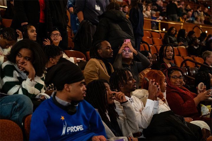 A group of students sit in the theater seats. Some converse with each other while others hold up their phones to take pictures.