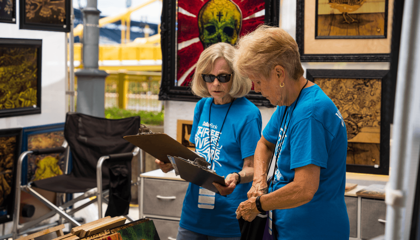 Ginny consults her clipboard as she stands beside another volunteer. Both are dressed in blue volunteer t-shirts with an artist market booth in the background.