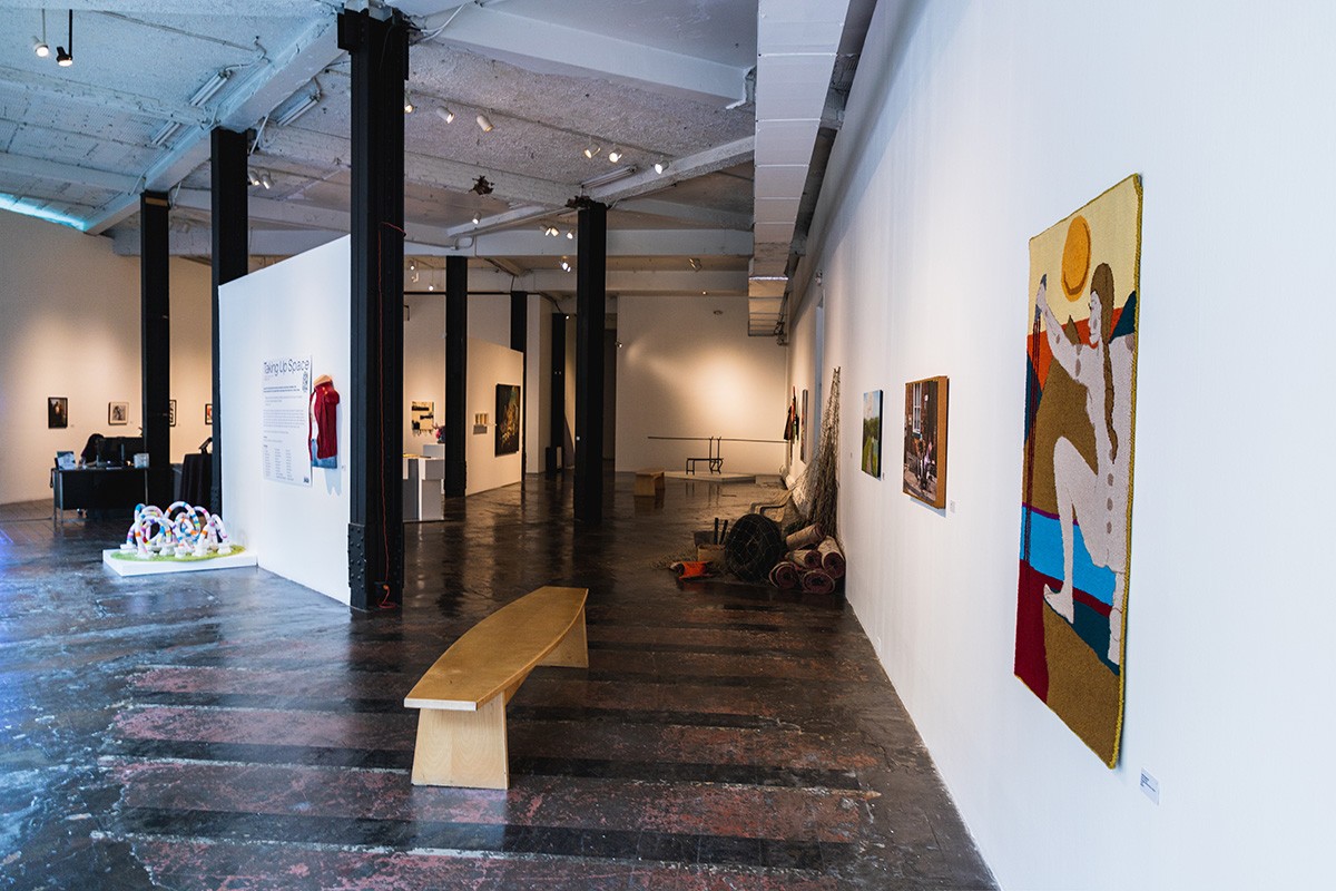 a large industrial gallery space filled with work hung on the walls and sitting on the floor. a bench sits on in the foreground.