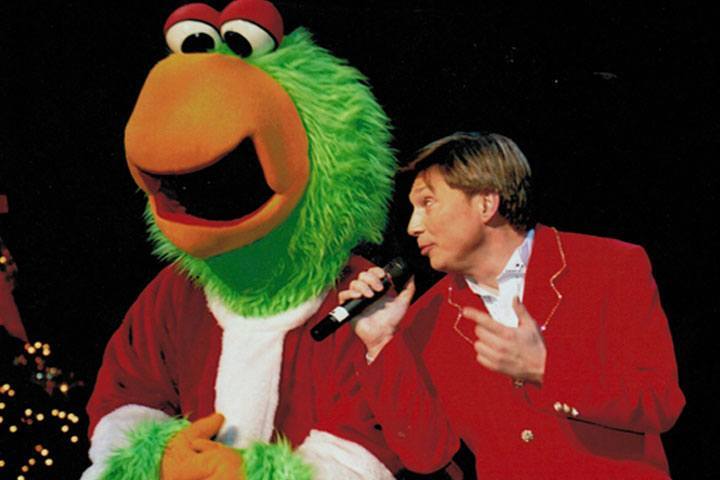 A man in a red suit jacket holds a microphone. He looks to the green Pittsburgh Pirates Pirate.