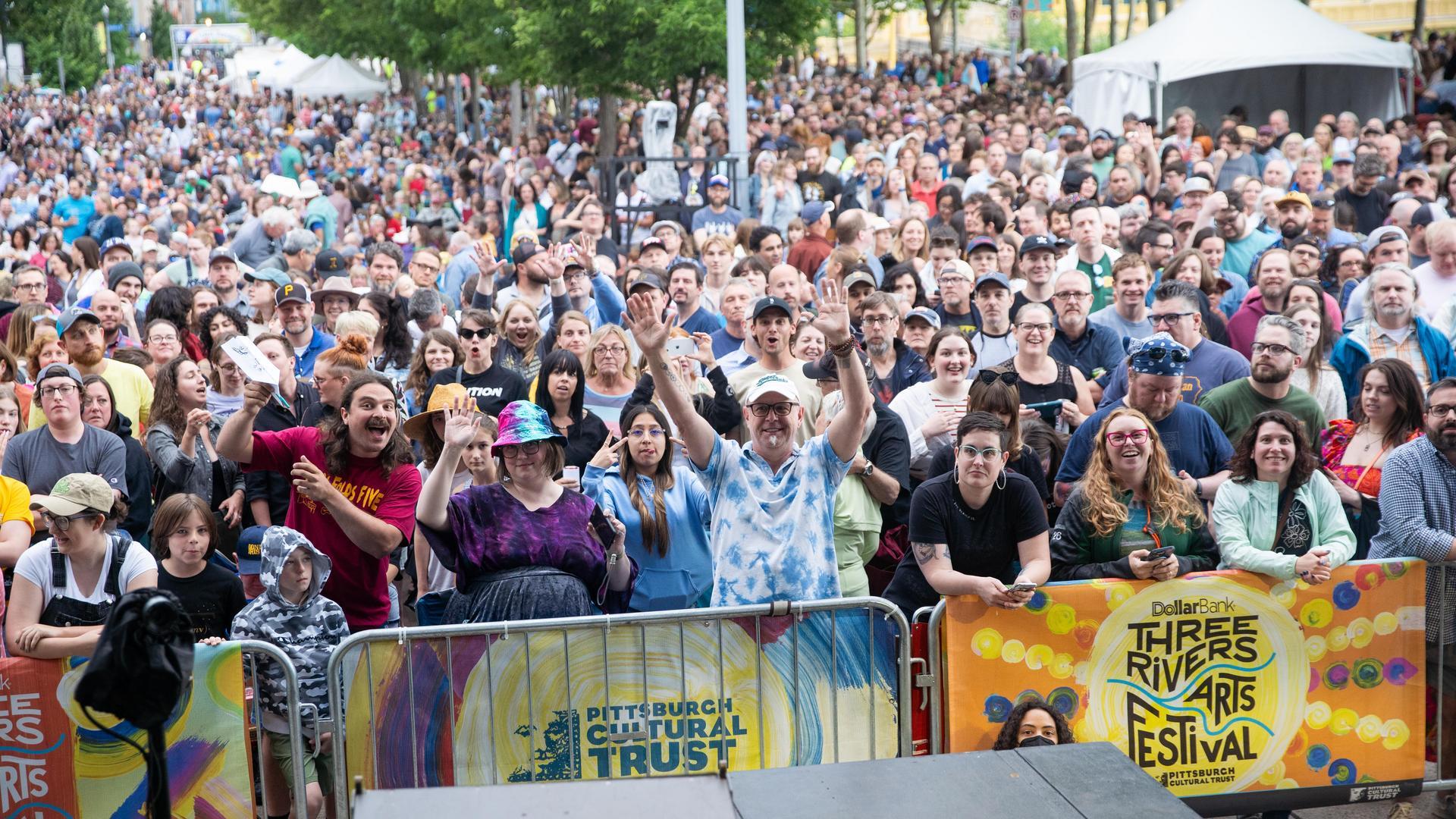 A large, diverse crowd stands outdoors behind barricades at the Three Rivers Arts Festival in Pittsburgh, smiling and waving at the camera on a bright day, with festival banners visible in front.