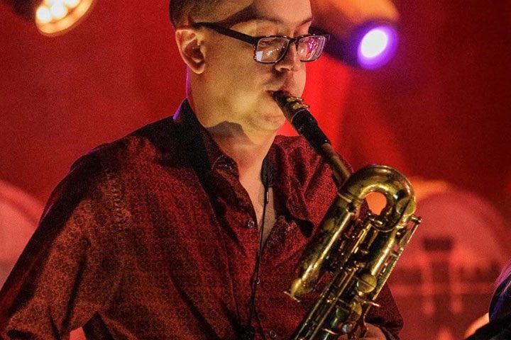 A man in glasses and a red button down shirt plays the saxophone.