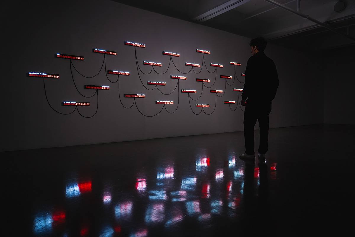 a large, dim gallery space. a white wall is filled with a spider web of small LED panels connected by wires. the panels display dozens of numerical statistics in red text. a silhouette of a person stands in front of the wall