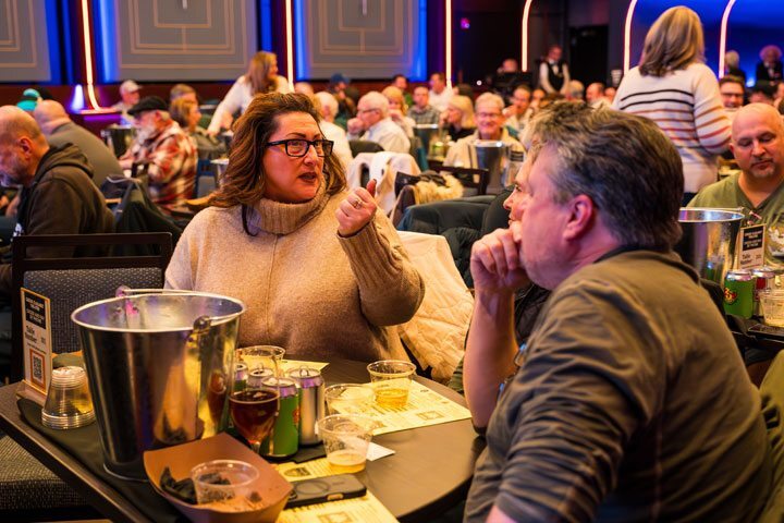 A woman and man, in mid conversation, sit at a table with cups and cans for beer in front of them. Behind them, several other people sit at tables.