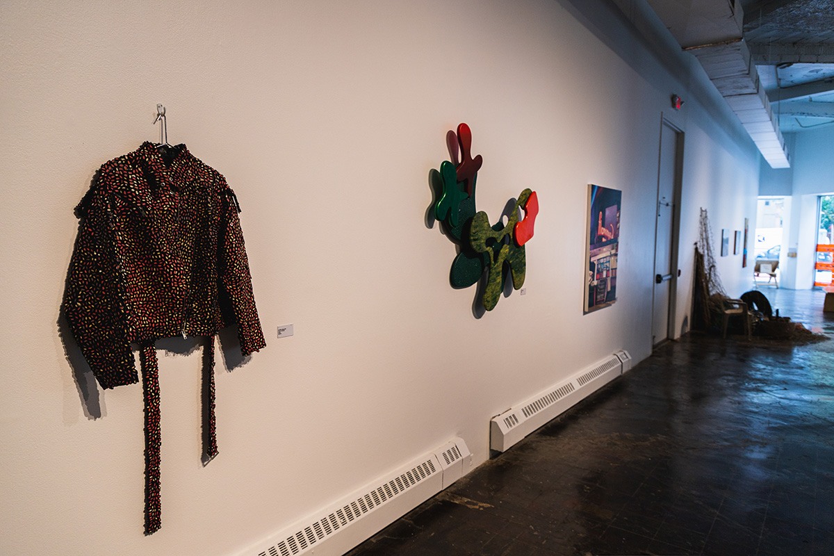 an ornamented jacket hangs on a white gallery wall. other pieces are out of focus in the background.