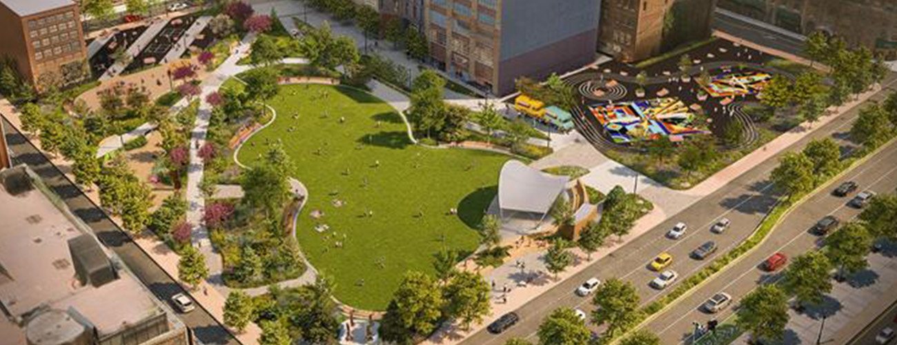 Aerial view of an urban park with a large green lawn, walking paths, scattered trees, colorful playground, and nearby buildings. People are relaxing on the grass and cars are driving on the adjacent road.