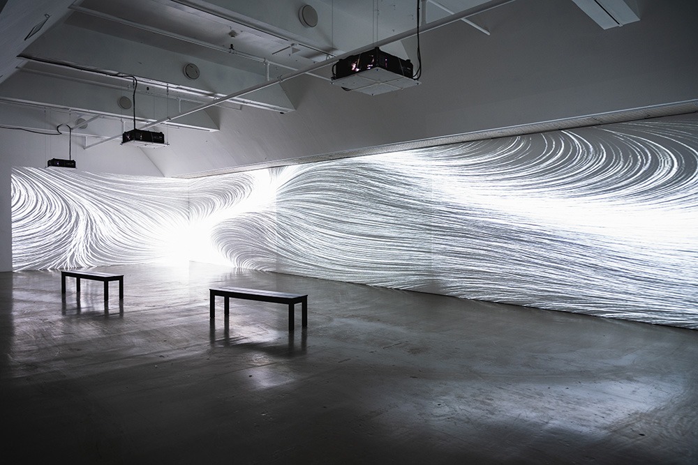 a large gallery space brightly lit by wave-like projections of white light on two walls. two benches sit in the middle of the room