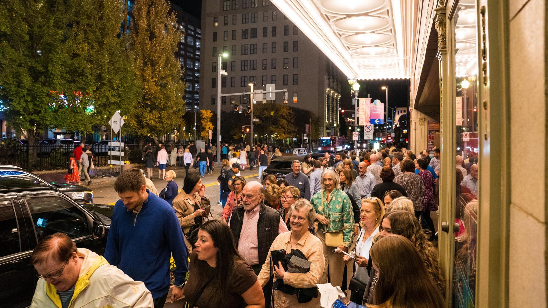 A large crowd of people stands in line outside a brightly lit theater at night, with trees, streetlights, and tall buildings visible in the background.