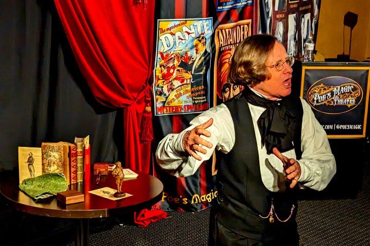 A man in glasses with a black cravat and blue waistcoat gestures to a crowd.