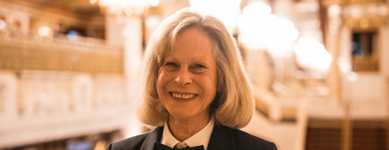 A woman in a formal black and white uniform smiles at the camera with the crystal chandeliers of the Bendedum Center lobby behind her.