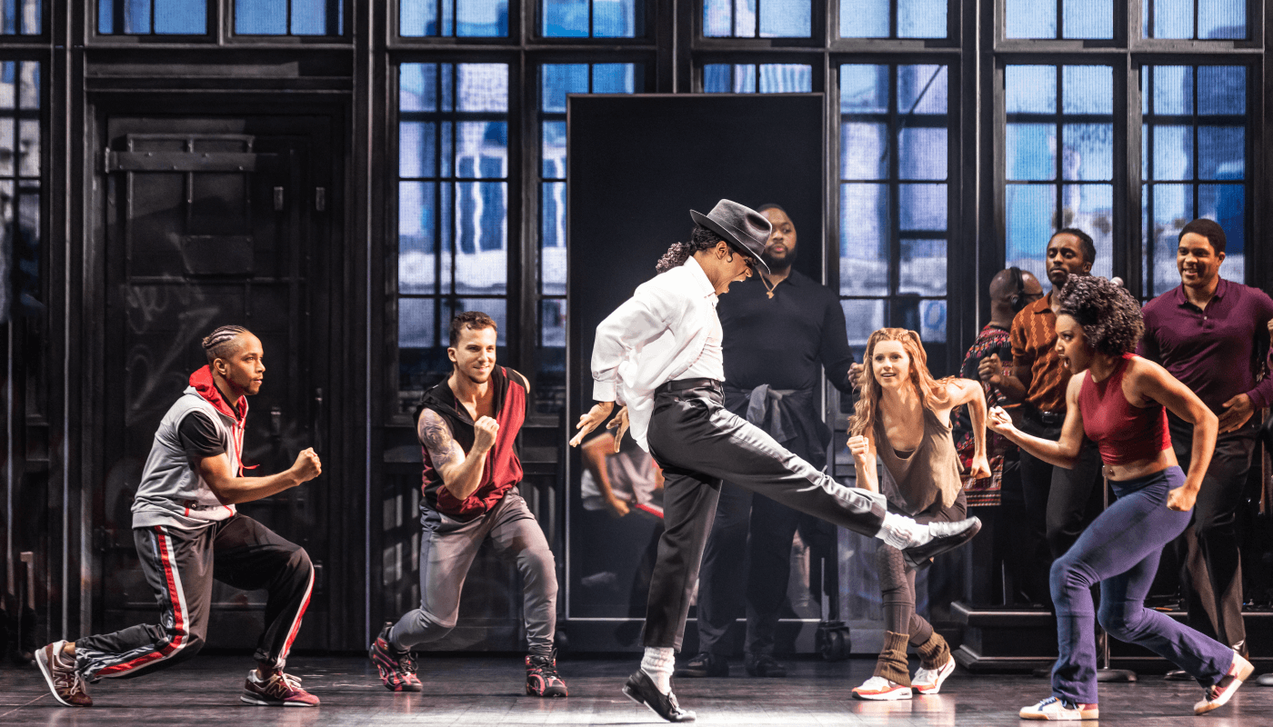 A man dressed as Michael Jackson and wearing a black fedora dances in front of a group of young actors.