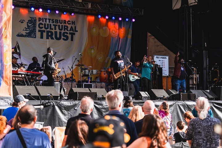 A group of musicians plays on an outdoor elevated stage with the Pittsburgh Cultural Trust logo behind them. A crowd of people stand in the foreground, slightly blurred.