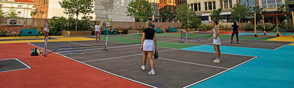 A group of people, mostly women, are playing pickleball on a colorful outdoor court in an urban setting. Several people watch from nearby seating areas.