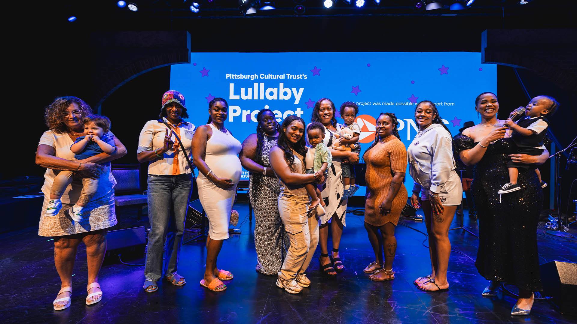 A group of adults standing on stage in front of a projection screen. Some of the adults are holding young kids.