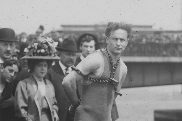 Black and white photo of Harry Houdini with chains wrapped around him and a crowd behind him