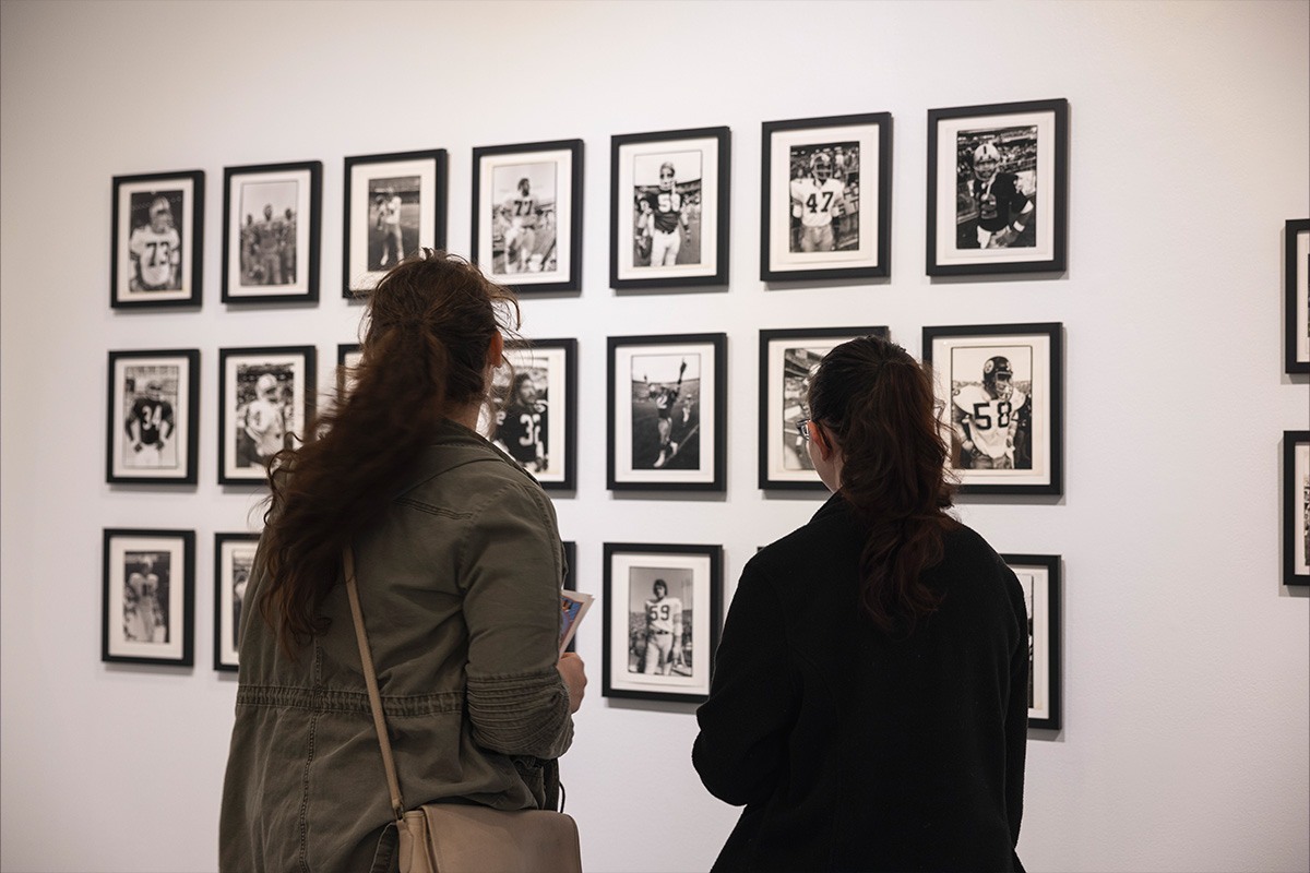 Two people stand in front of a white wall displaying a grid of framed black-and-white photographs, viewing and discussing the images in an art gallery or museum setting.