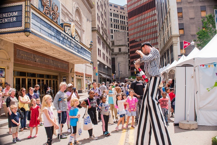 A man dressed in black and white stripes walking on tall stilts entertains a group of children outside the Benedum Center.