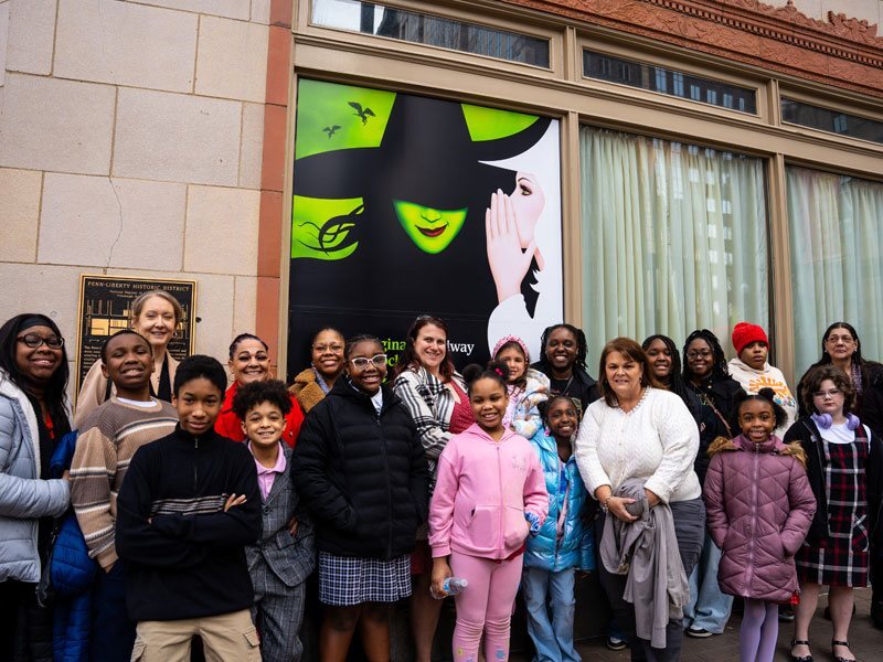 Students and teachers stand in front of the theater with a Wicked poster behind them.