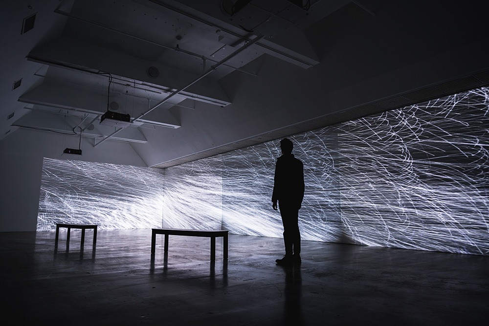 a large, dim gallery space with white lines projected on two walls. two benches and a person standing are silhouetted in the middle of the space