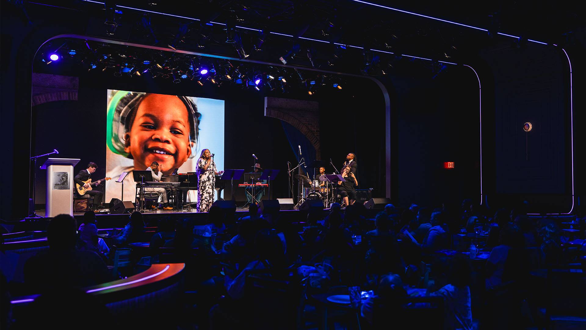 A band on a stage performing music. Behind them is a projection of a child's face. A audience watches in a dark hall.