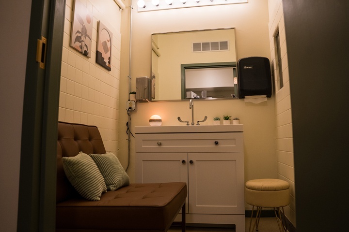 Taken from the entrance of the lactation room, you can see a chocolate brown lounge chair with two green pillows, a cushioned stool, and a white vanity with a silver faucet. The walls are a warm white color and botanical canvas prints are hung on the wall.