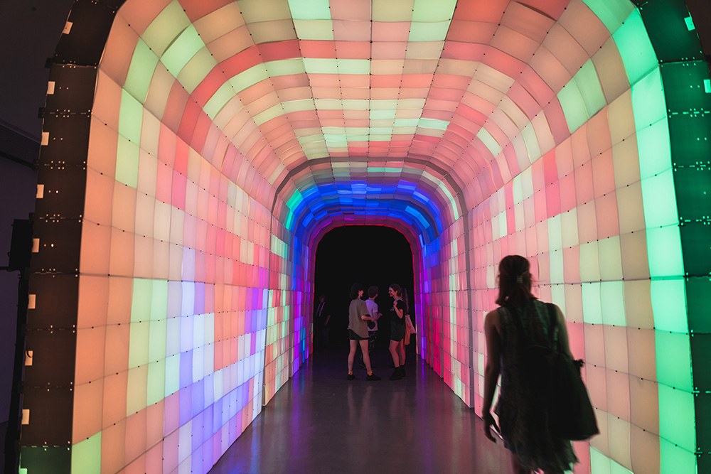 people walking through a wide, arched tunnel made of multicolored light up blocks