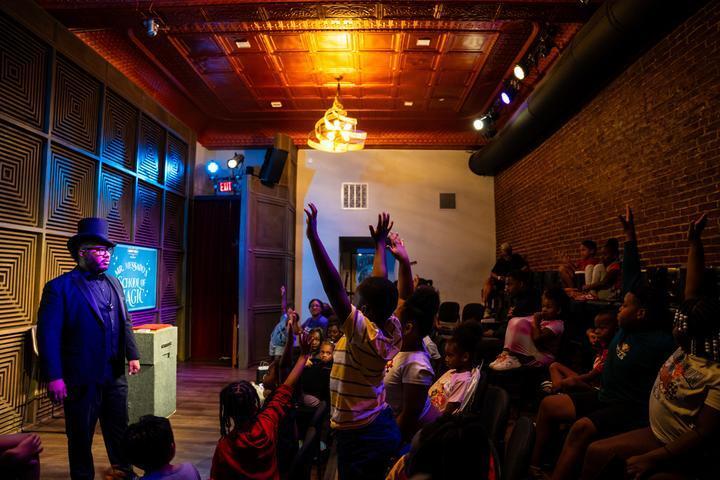 Messado looks out on the crowd as children raise their hands, some are standing up.