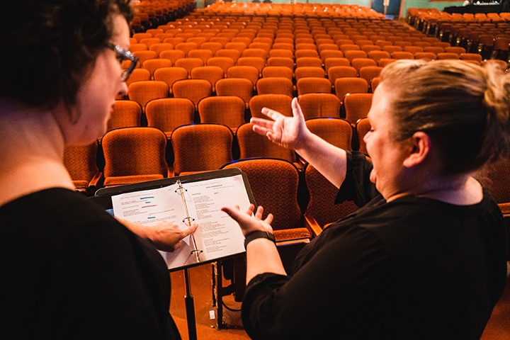 a view over the shoulders of two white women in black clothing. the woman on the left points at a printed script on a music stand while the woman on the right makes a sign