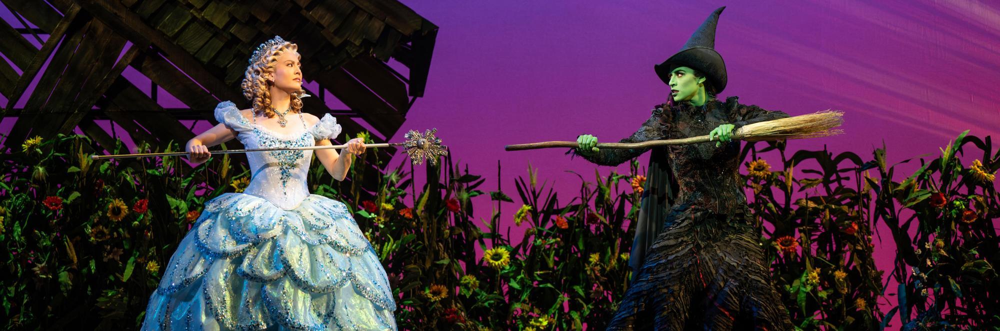 Two women on stage in elaborate costumes—one in a sparkling blue gown holding a wand, the other in a black dress, green face paint, and a pointy hat holding a broom—stand facing each other against a colorful backdrop.