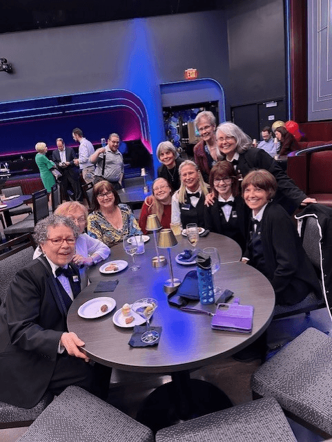 A group of volunteers in their uniform black jackets and white button downs sit around two tables at the Greer Cabaret Theater.