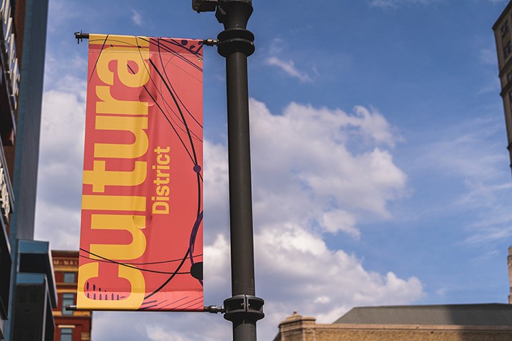 an orange and yellow banner hangs from a light pole in front of a blue sky dotted with clouds.