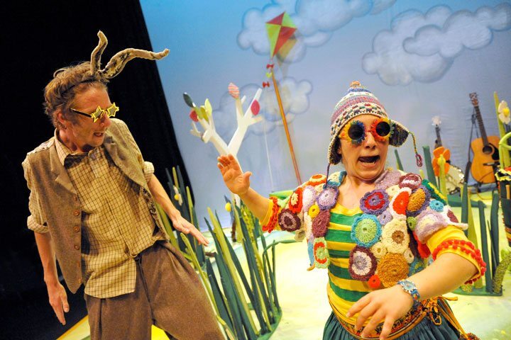 A woman with multi-colored, rounded sunglasses and a crocheted vest turns her head off stage. A man with star-shaped sunglasses and a headband with horns attached look down and to the side. Behind them in a painted sky background and long grasses made of wood.