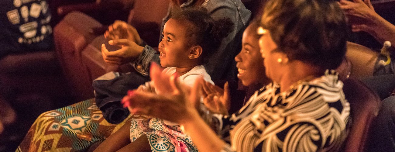 an adult woman in the theater holds up a program and looks over the shoulder of the young girl sitting next to her.