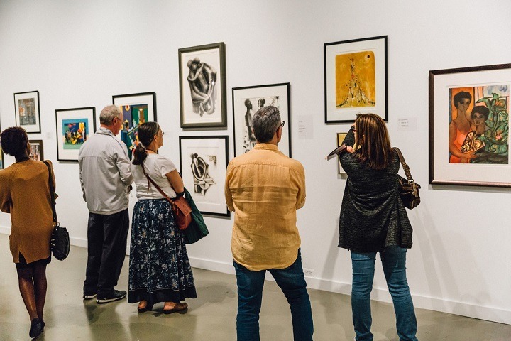 Two pairs of people and one individual gaze at several framed pieces of artwork hung on a white gallery wall.