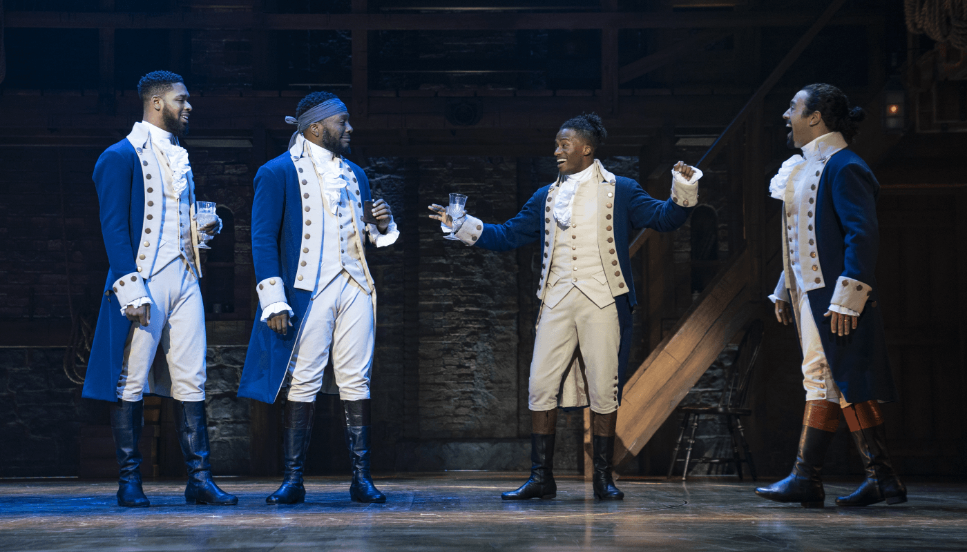 a group of four men stand on stage, listening to one of these people talking animately. They are dressed in 18th century garb of long blue jackets white ruffled shirt