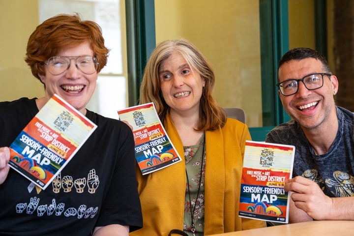 Two women and a man smile while sitting at a conference table and holding up three sensory friendly maps.