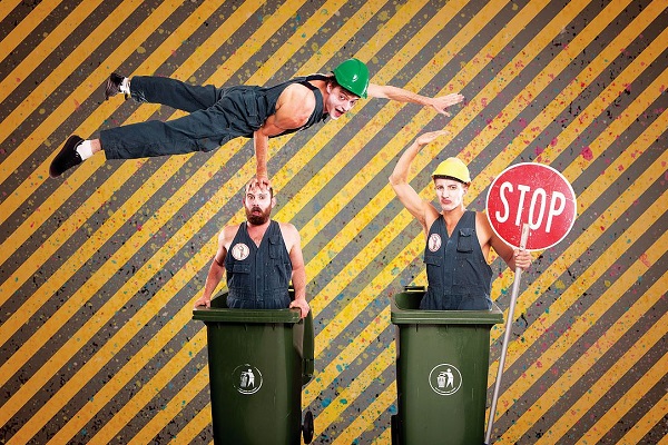 One man balancing on top of a man in a trash bin. Another man in trash bin holding a stop sign