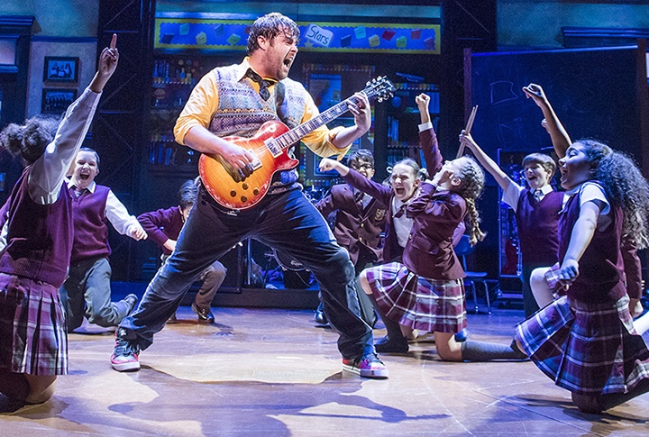 a man plays guitar in the center of the photo; around him, a group of students kneel on the floor with their fists in the air