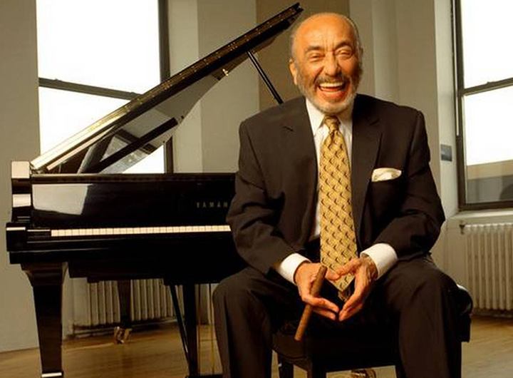 Eddie Palmieri sits on a bench in front of a piano wearing a suit and cheery yellow tie