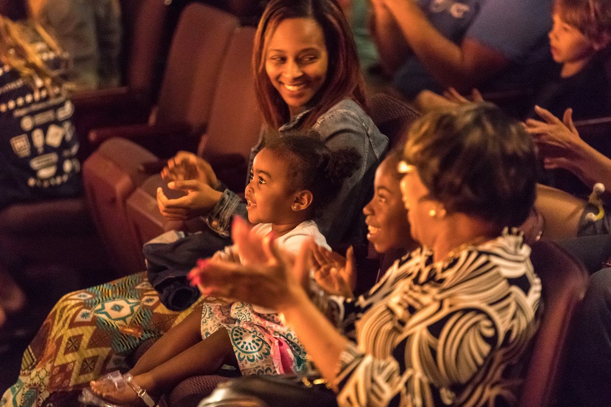 a pre-school aged child claps while her family smiles beside her