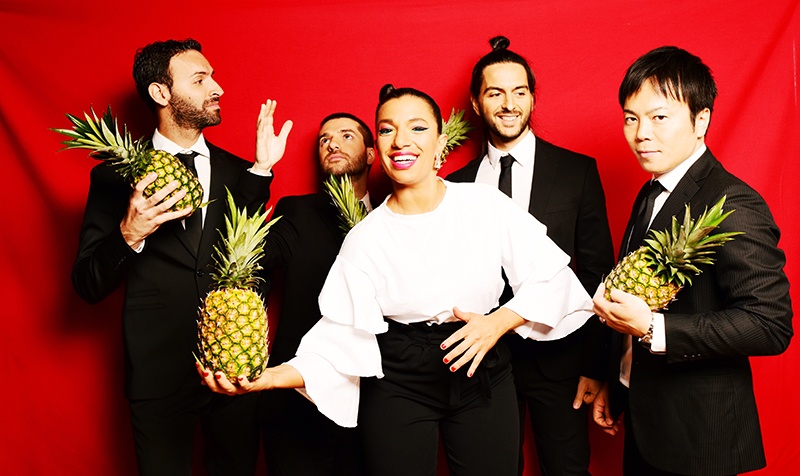 woman in white shirt holding a pineapple, four people behind her, laughing