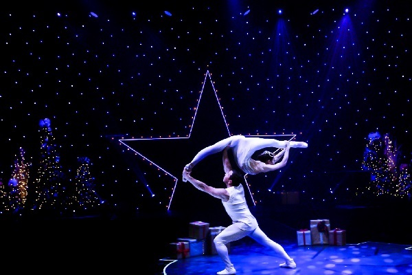 one circus performer holding another in front of a lit up star outline surrounded by Christmas trees
