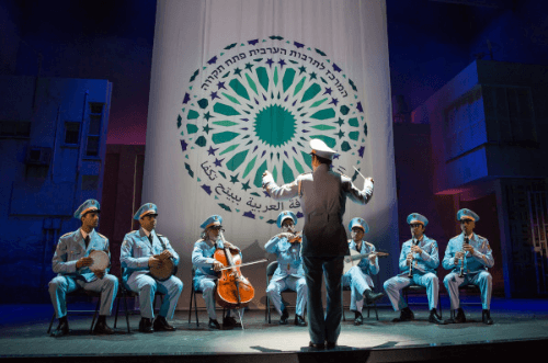 A man conducts a small band, sitting and playing their instruments, dressed in blue uniforms