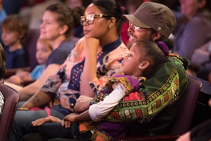 a young child sits in their parent's lap as they watch a show in a theater