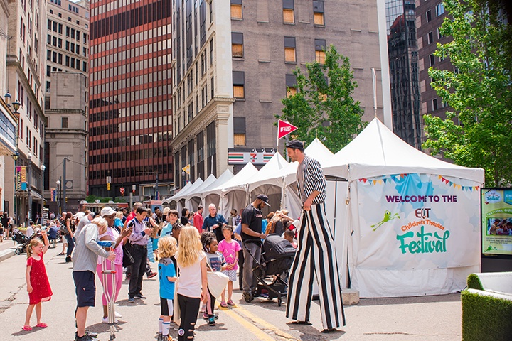 a crowd of children and parents being entertained by a man on stilts at the 2018 eqt children's theater festival