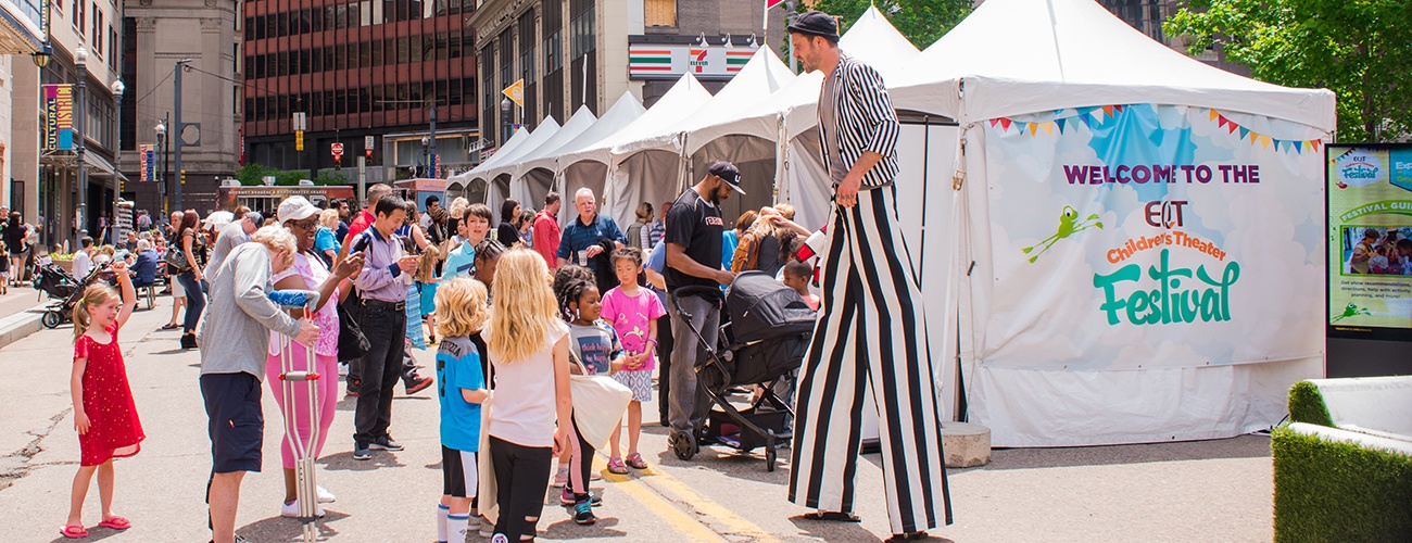 a crowd of children and parents being entertained by a man on stilts at the 2018 eqt children's theater festival