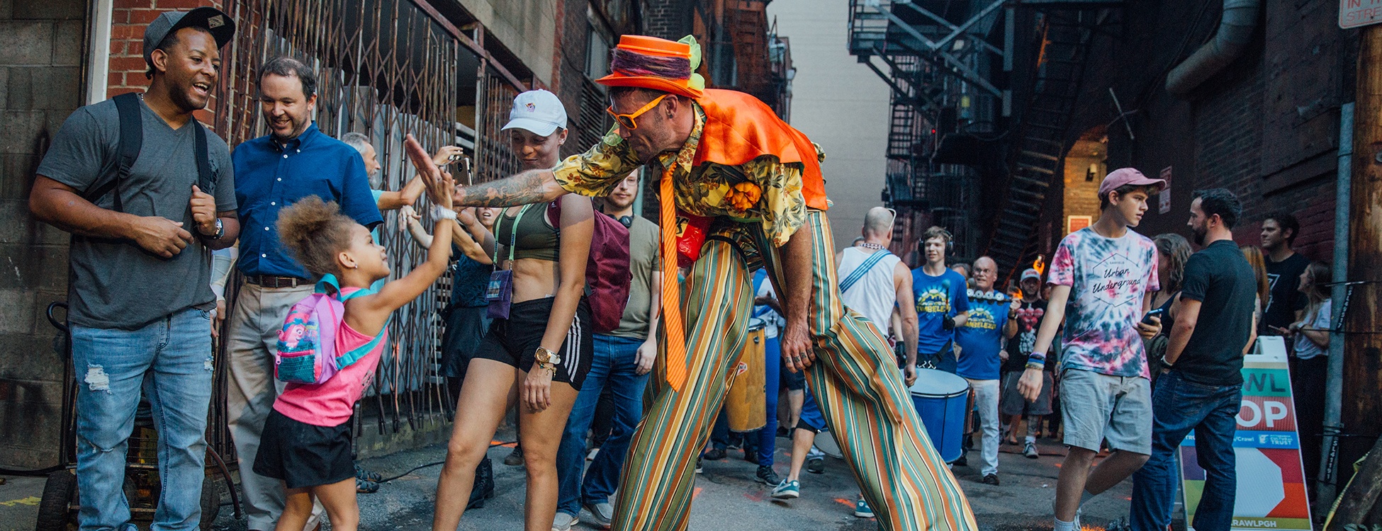a performer on stilts gives a high five to a young child at a previous art in the alley event