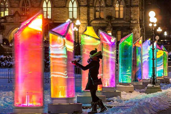 woman dressed in coat, hat, and snow boots touching prism of colorful light installation Prismatica