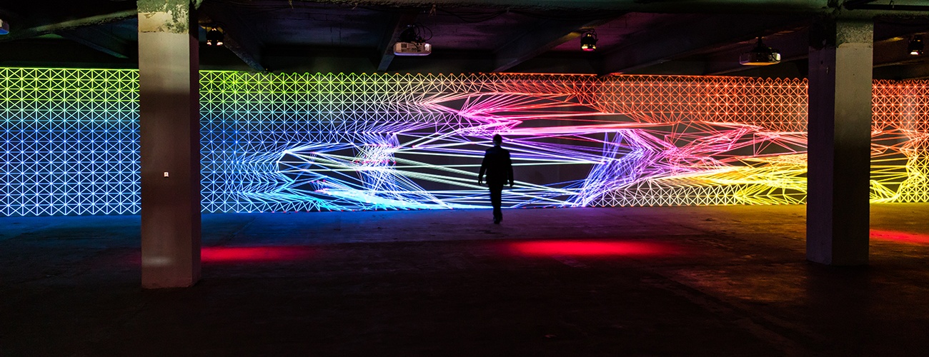 a person stands in a large, open room, viewing a light installation being projected on the far wall